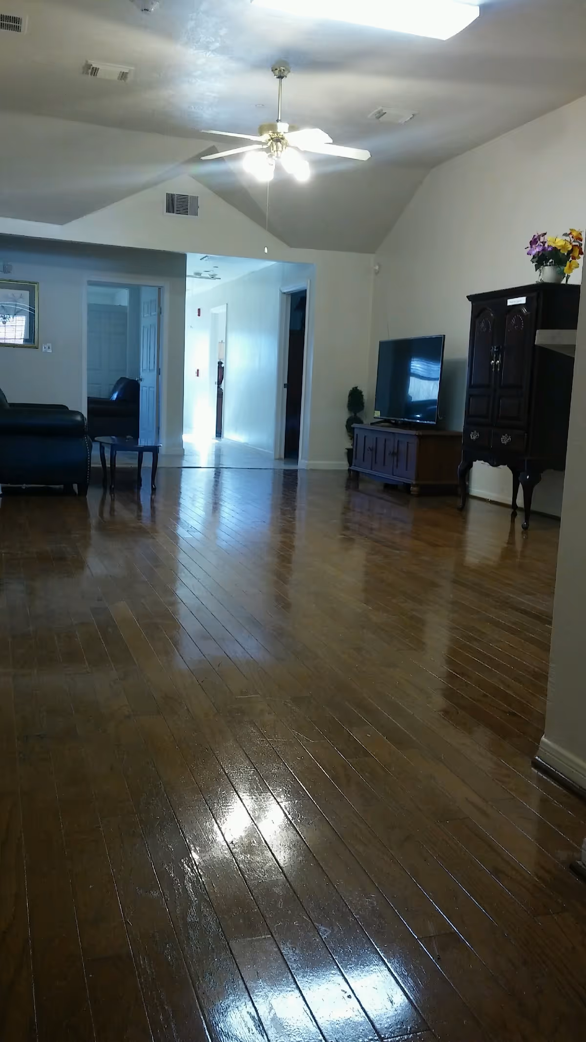 Interior view of a living room with polished wooden floors, a ceiling fan with lights, a black leather chair, a wooden TV stand with a flat-screen television, and a dark wooden cabinet with flowers on top. The room has white walls and an open doorway leading to a hallway.