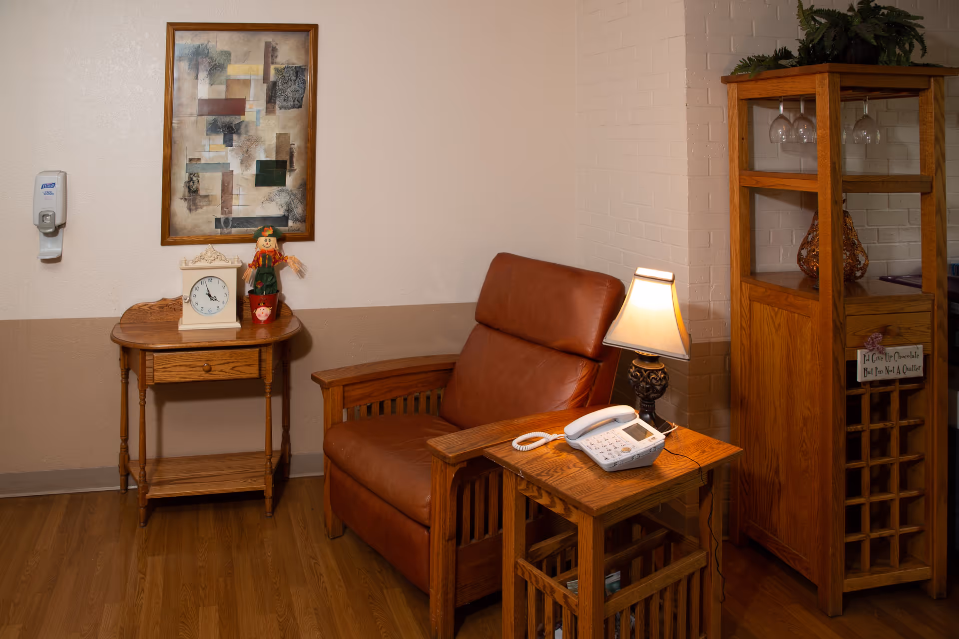 A cozy corner of a room featuring a brown leather armchair next to a wooden side table with a lamp and a white telephone. To the left, there is a small wooden table with a clock and a decorative scarecrow figure. Above the table hangs an abstract framed painting. On the right side, there is a wooden cabinet with glassware and decorative items. The walls are painted beige with a darker beige lower half, and the floor is wooden.