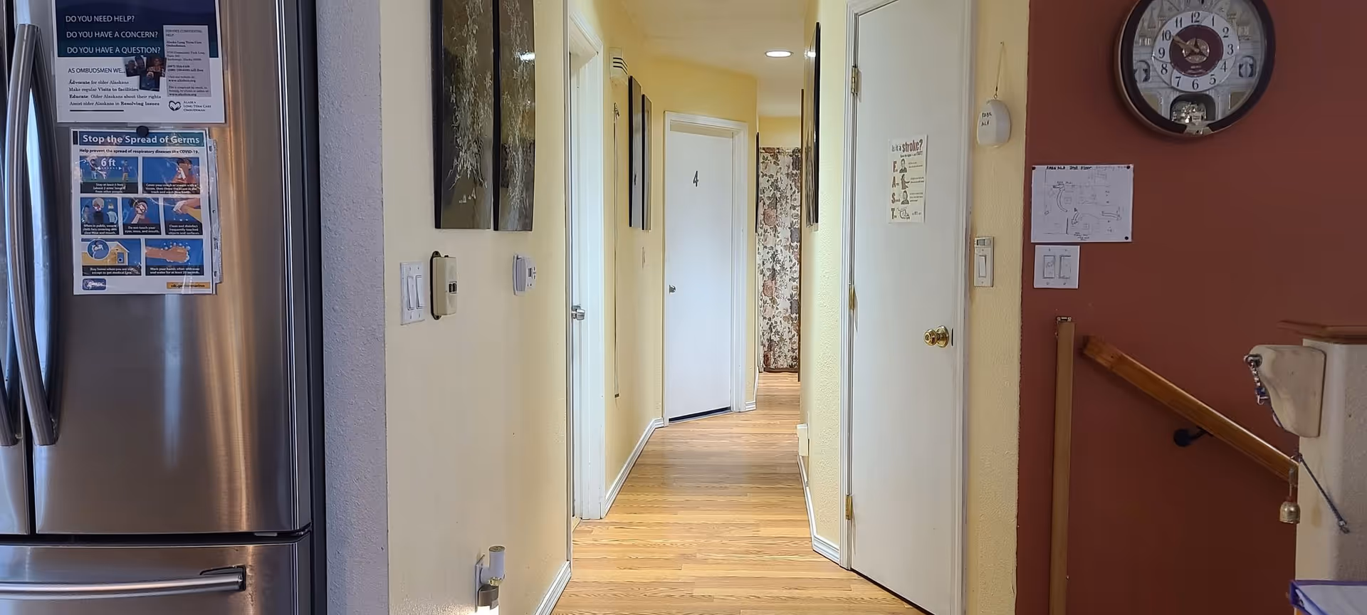 Bright hallway inside a senior living facility with doors, clock, and stainless steel refrigerator.