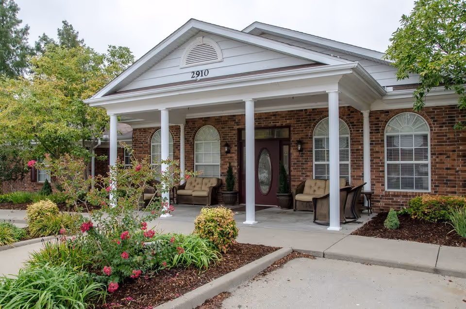 Front entrance of a brick senior living building with a covered porch supported by white columns, outdoor seating, and landscaped flowerbeds.