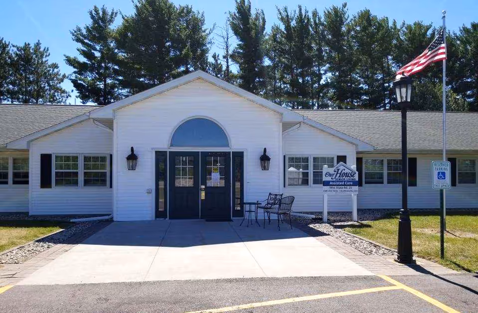 Front exterior of a single-story white senior living building with a central entrance, double doors, a sign, flagpole, and outdoor seating.