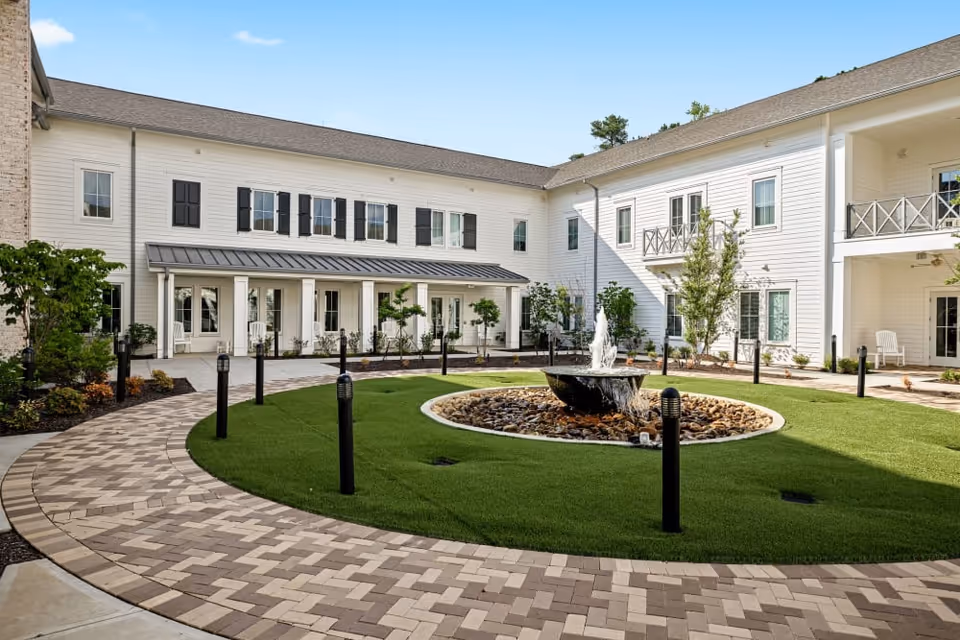 Outdoor courtyard area of The Claiborne at Shoe Creek featuring a circular water fountain surrounded by green grass and a paved walkway. The courtyard is enclosed by a two-story white building with multiple windows, balconies, and a covered porch with seating.