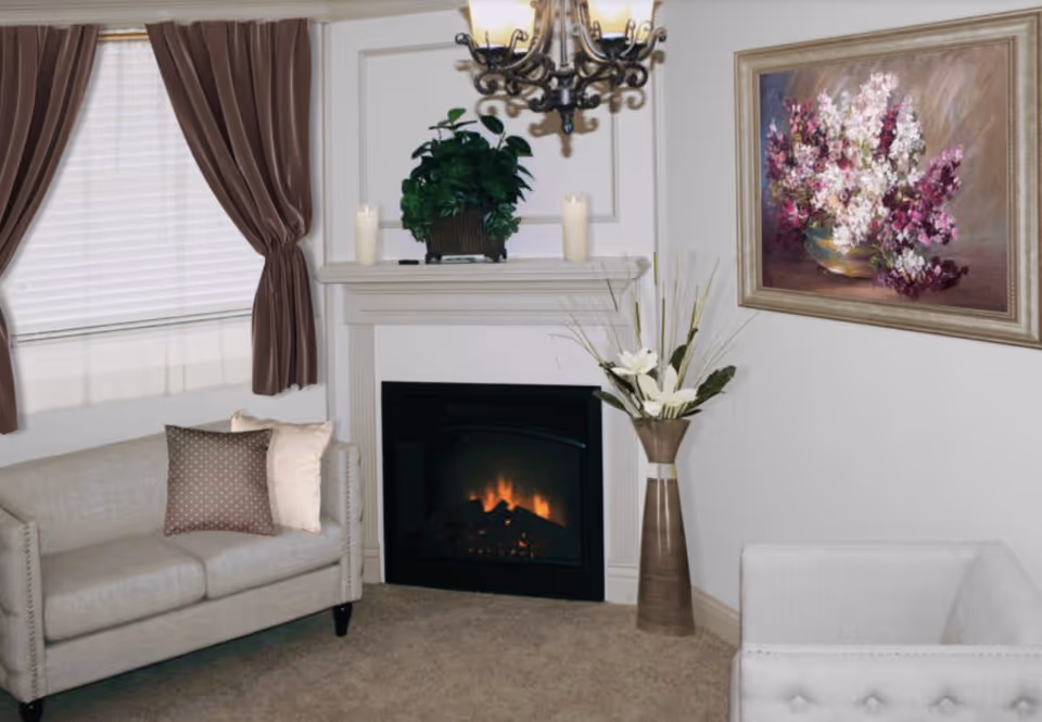A cozy living room with a beige sofa adorned with two pillows, a fireplace with a fire burning, a plant and two candles on the mantel, a tall vase with white flowers, brown curtains on the window, and a framed painting of purple and white flowers on the wall.