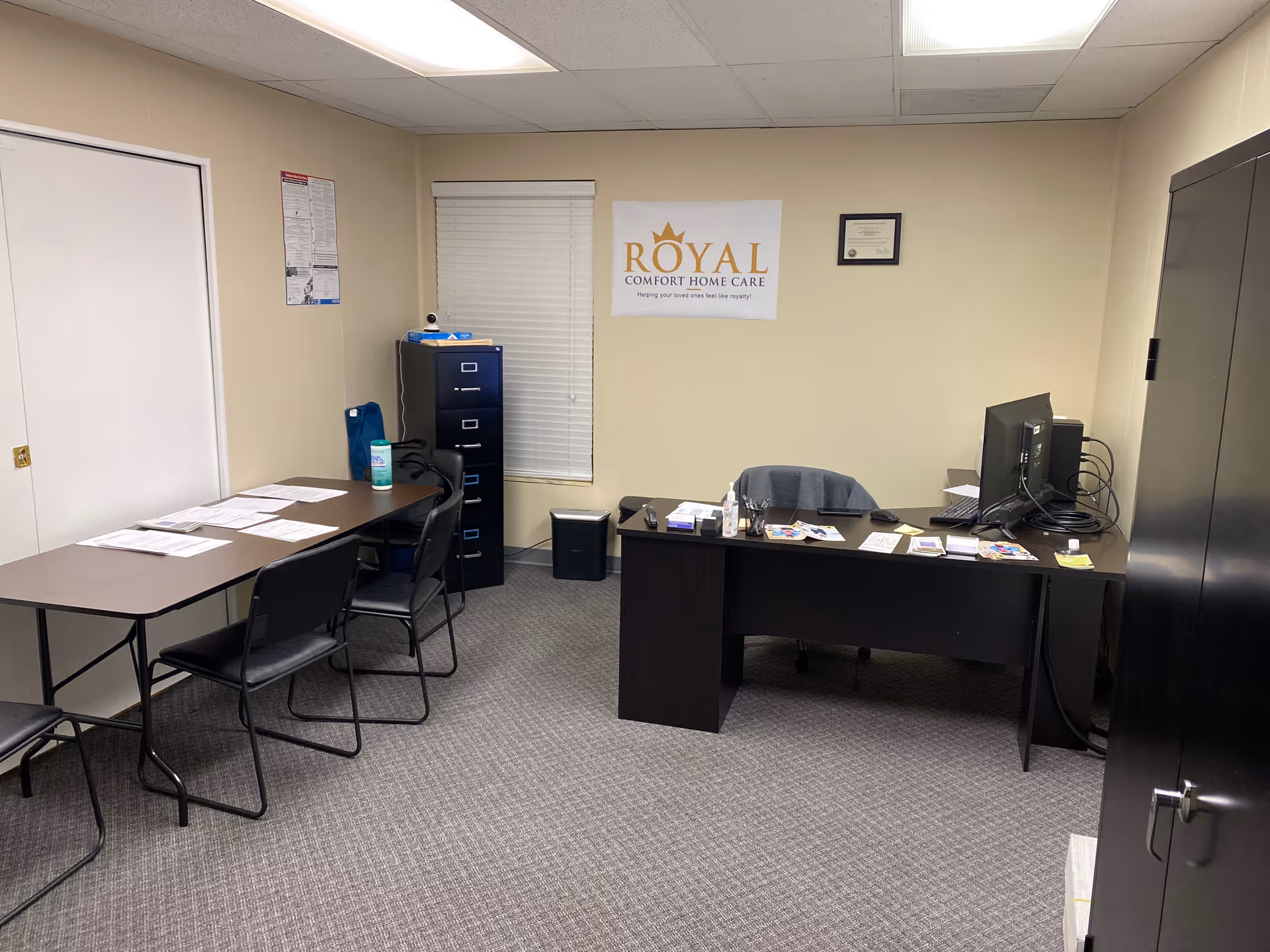 Office space at Royal Comfort Home Care with desk, chairs, and banner on wall.