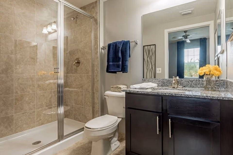 Clean modern bathroom with glass-enclosed shower, toilet, and dark vanity with granite countertop and mirror.