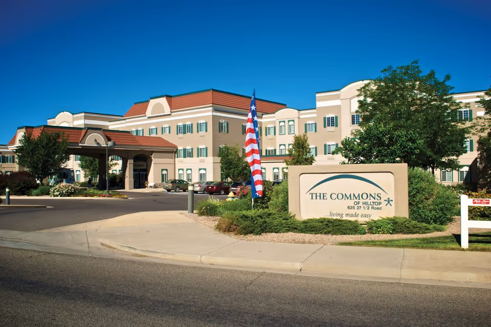 Front exterior of The Commons of Hilltop senior living building with a porte-cochere, landscaped grounds, American flag, and a stone entrance sign.