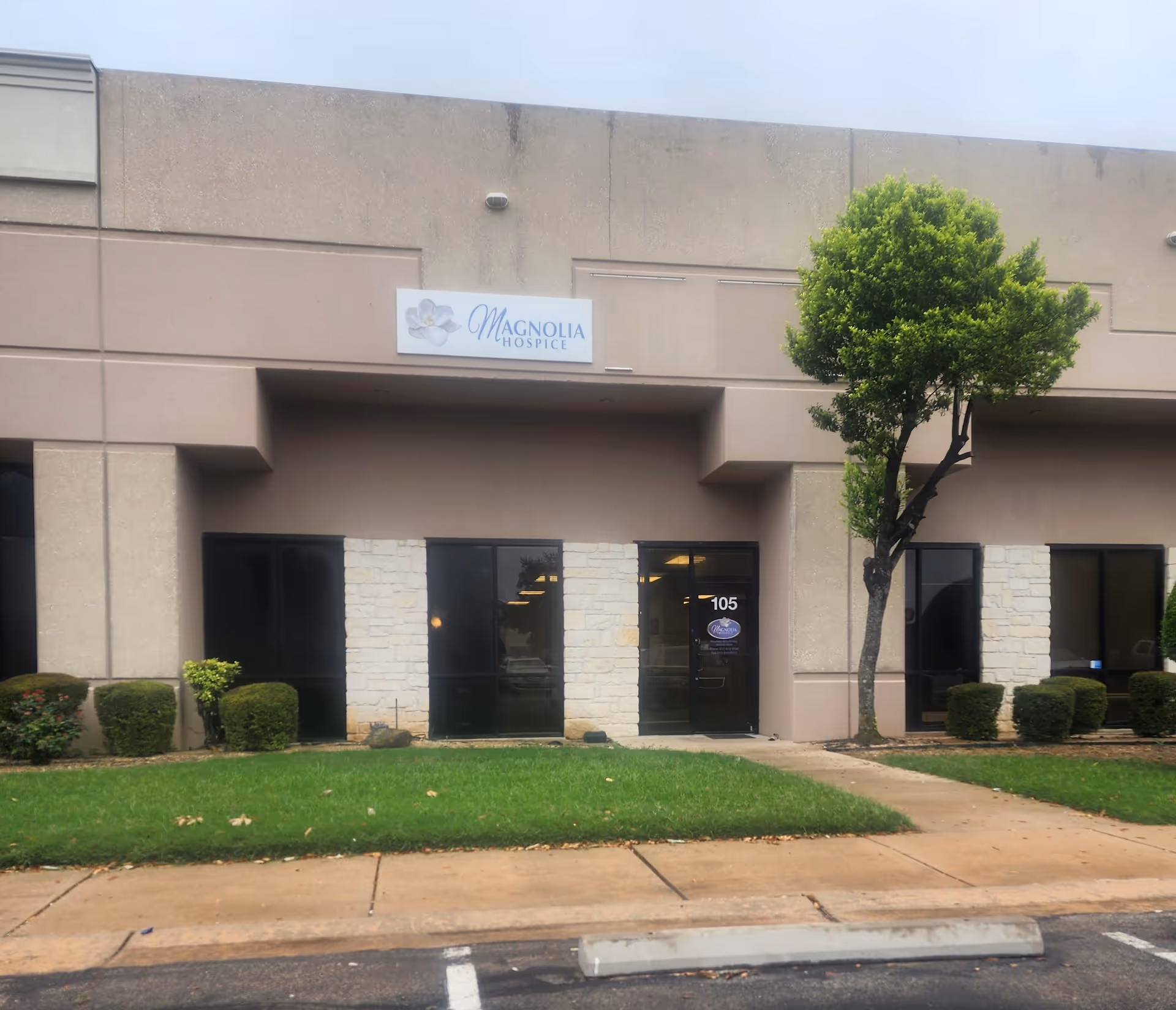 Exterior view of Magnolia Hospice building with entrance, glass doors, tree, and landscaping.