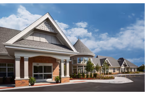 Exterior view of a senior living facility with brick entrance, gabled roof, and parking.