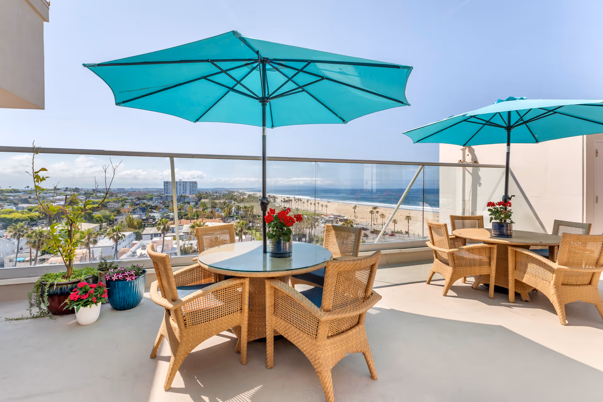 Outdoor patio area with two round glass-top tables surrounded by wicker chairs, each table shaded by a large turquoise umbrella. Potted plants with flowers are placed near the glass railing, which overlooks a beach, ocean, and distant cityscape under a clear blue sky.