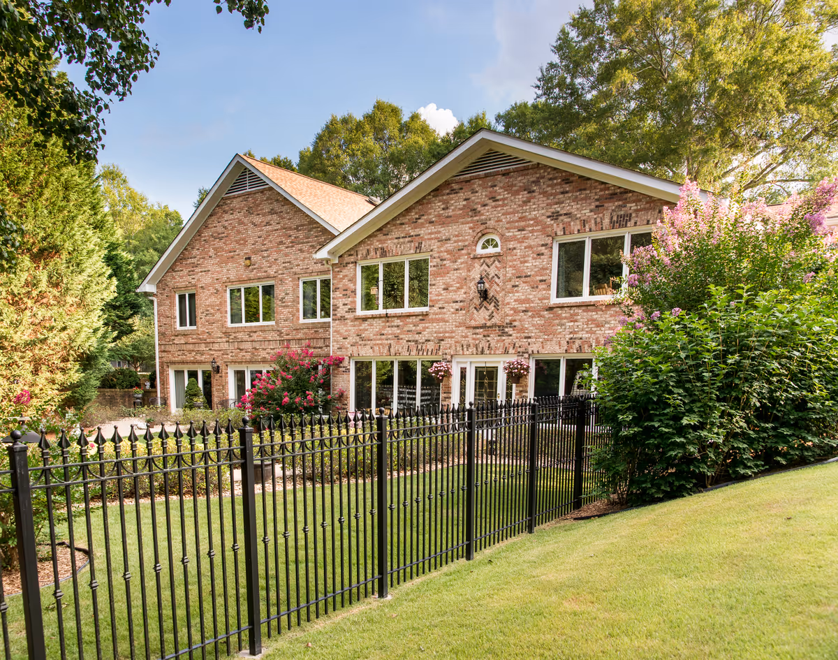 Exterior of Mayfield Oaks senior living building with brick facade, windows, black fence, and landscaping.