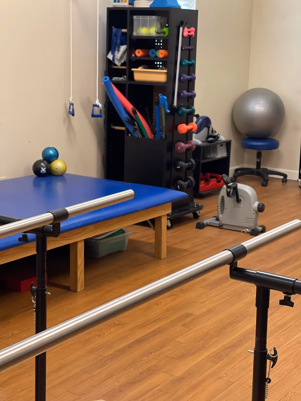 A therapy room with parallel bars, exercise equipment, a blue padded table, and a wooden floor.