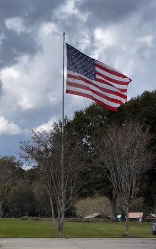 American flag waving atop a tall flagpole beside trees under cloudy sky.