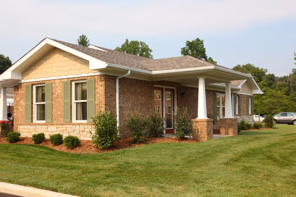 Exterior view of a single-story brick building with green shutters and a covered porch supported by white columns. The building is surrounded by well-maintained grass, shrubs, and trees, with a few parked cars visible in the background under a clear sky.
