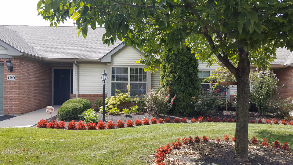 Exterior view of a single-story residential building with a brick and siding facade, a dark front door, and windows. The front yard features a well-maintained lawn, a tree, flower beds with red and green plants, and a lamp post near the walkway leading to the entrance. A security alarm sign is visible near the walkway.