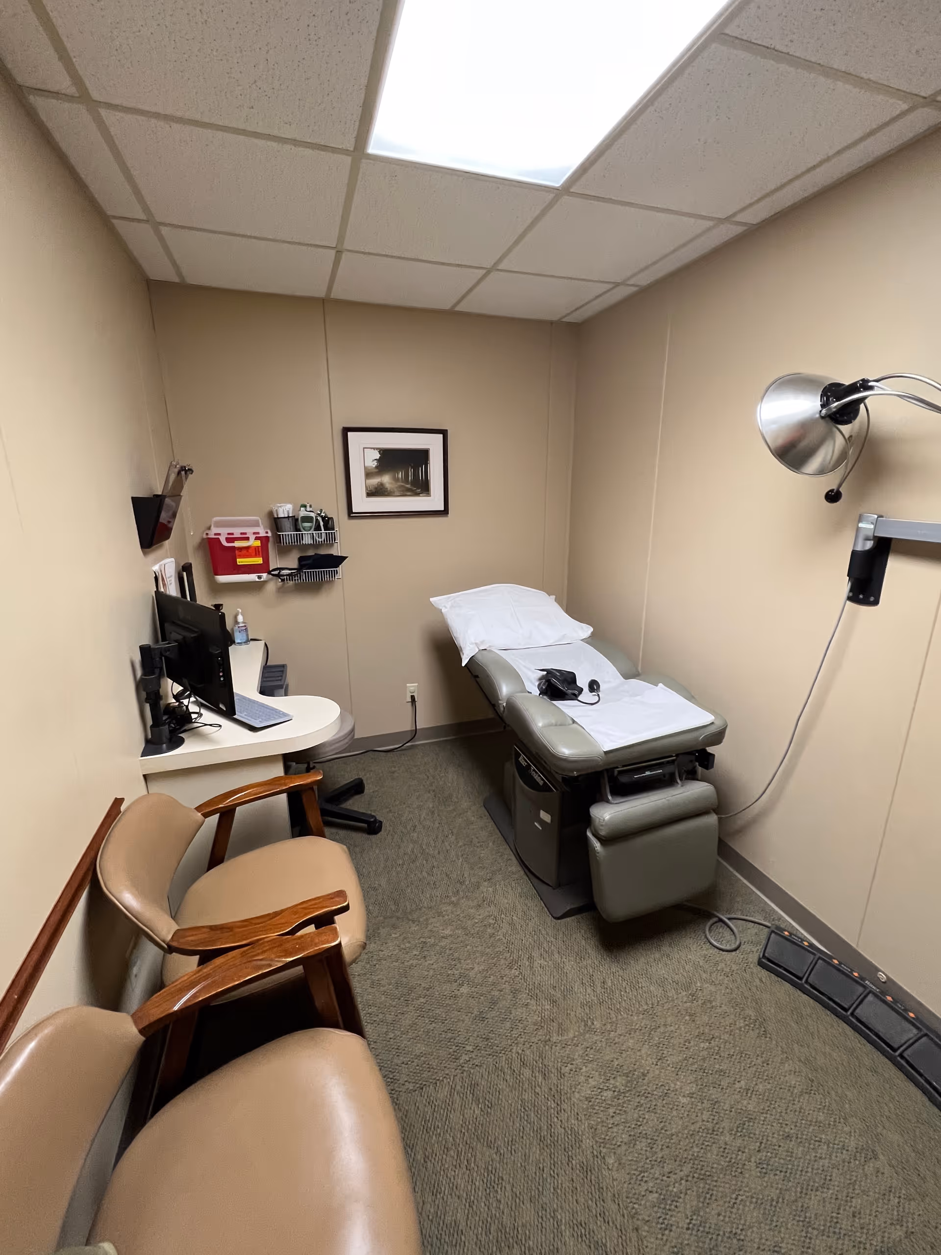 A small medical treatment room featuring an exam chair, computer station, and wall-mounted lamp.