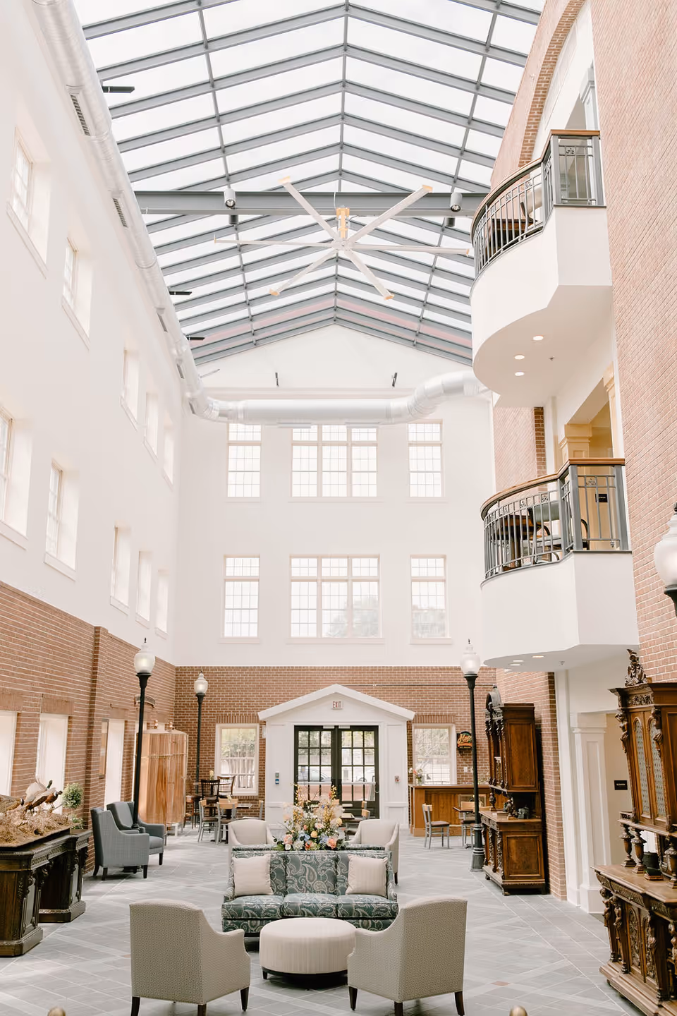 Bright multi-story atrium with a glass skylight, brick walls, balconies and a furnished seating area centered around a floral arrangement.