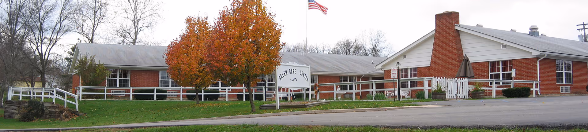 Front view of a single-story red-brick senior care building labeled 'Salem Care Center' with white railings, an American flag, and autumn trees.