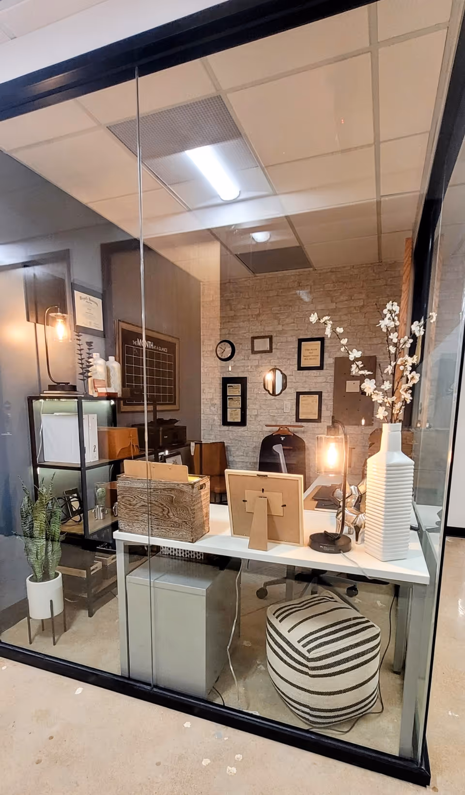Glass-walled reception area with desk, computer, decor, lamp, plant, and striped pouf.