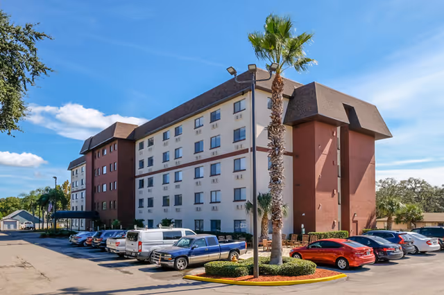 Five-story beige-and-red building with a parking lot, parked cars, and palm trees under a blue sky.