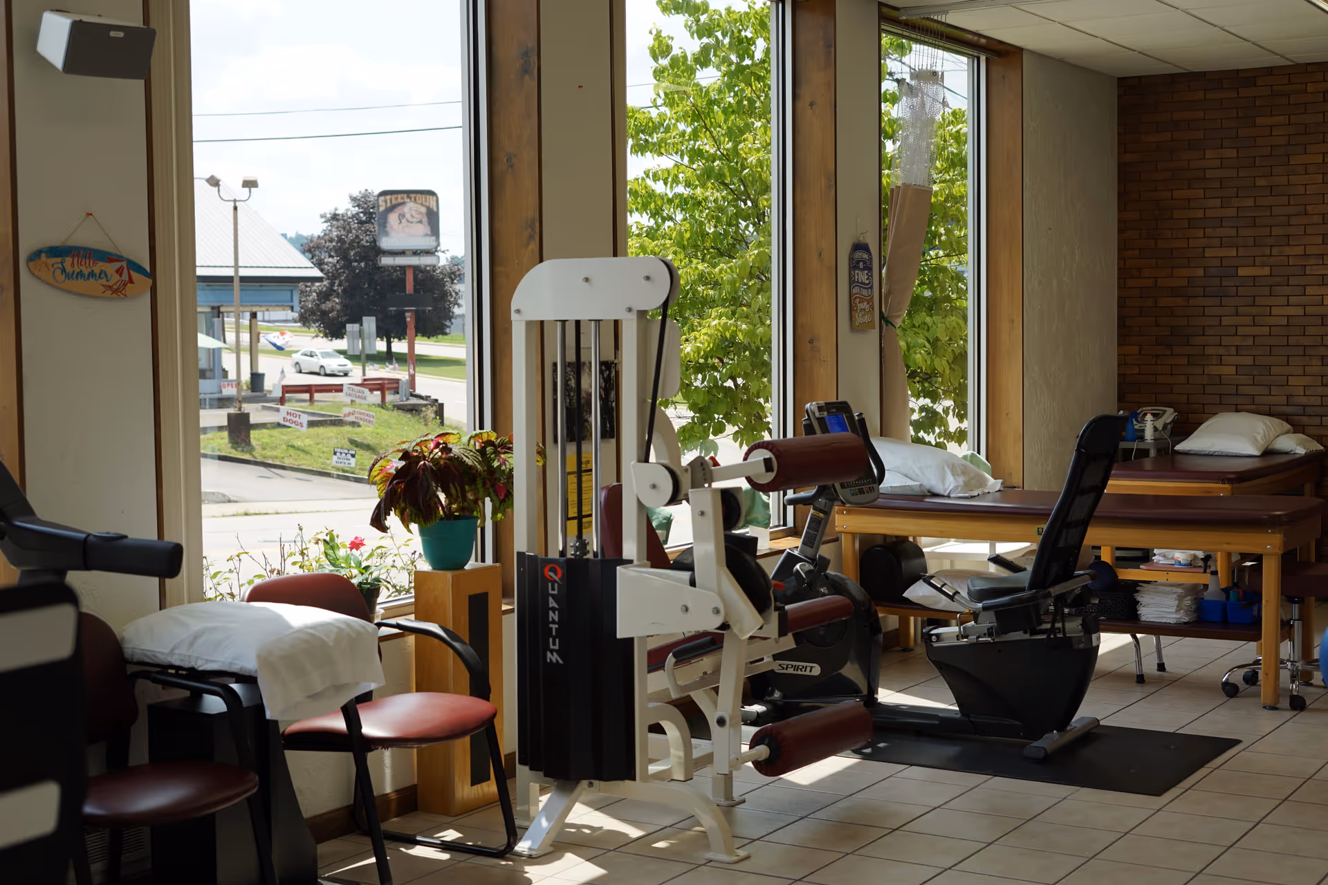 Interior physical therapy room with exercise machines, therapy tables, chairs, and large windows.