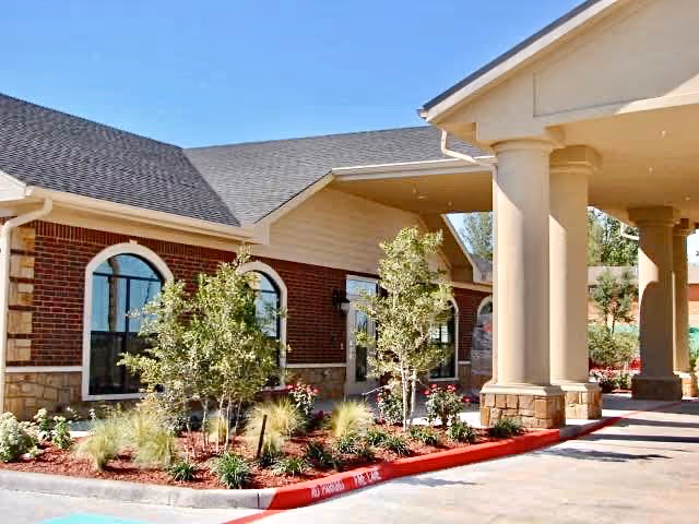 Exterior view of a building with brick and stone facade, arched windows, and a covered entrance supported by large columns. There are landscaped plants and small trees along the building with a clear blue sky above.