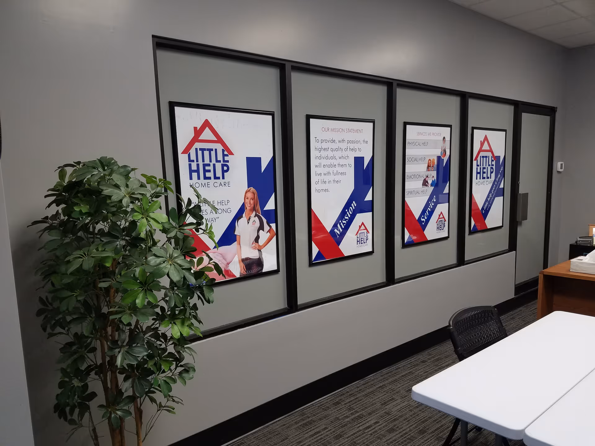 Indoor office hallway with framed Little Help Home Care posters and a potted plant.