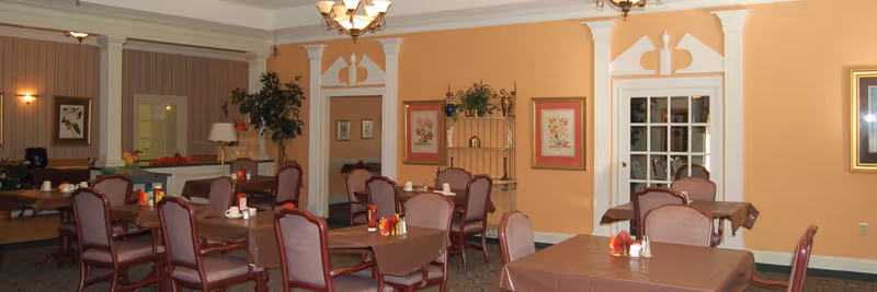 A dining room with several tables covered in brown tablecloths, each set with cups, napkins, and condiments. The room has peach-colored walls, framed floral artwork, a decorative shelving unit with plants, and a chandelier hanging from the ceiling. There are multiple chairs around the tables and a large plant near the back wall.