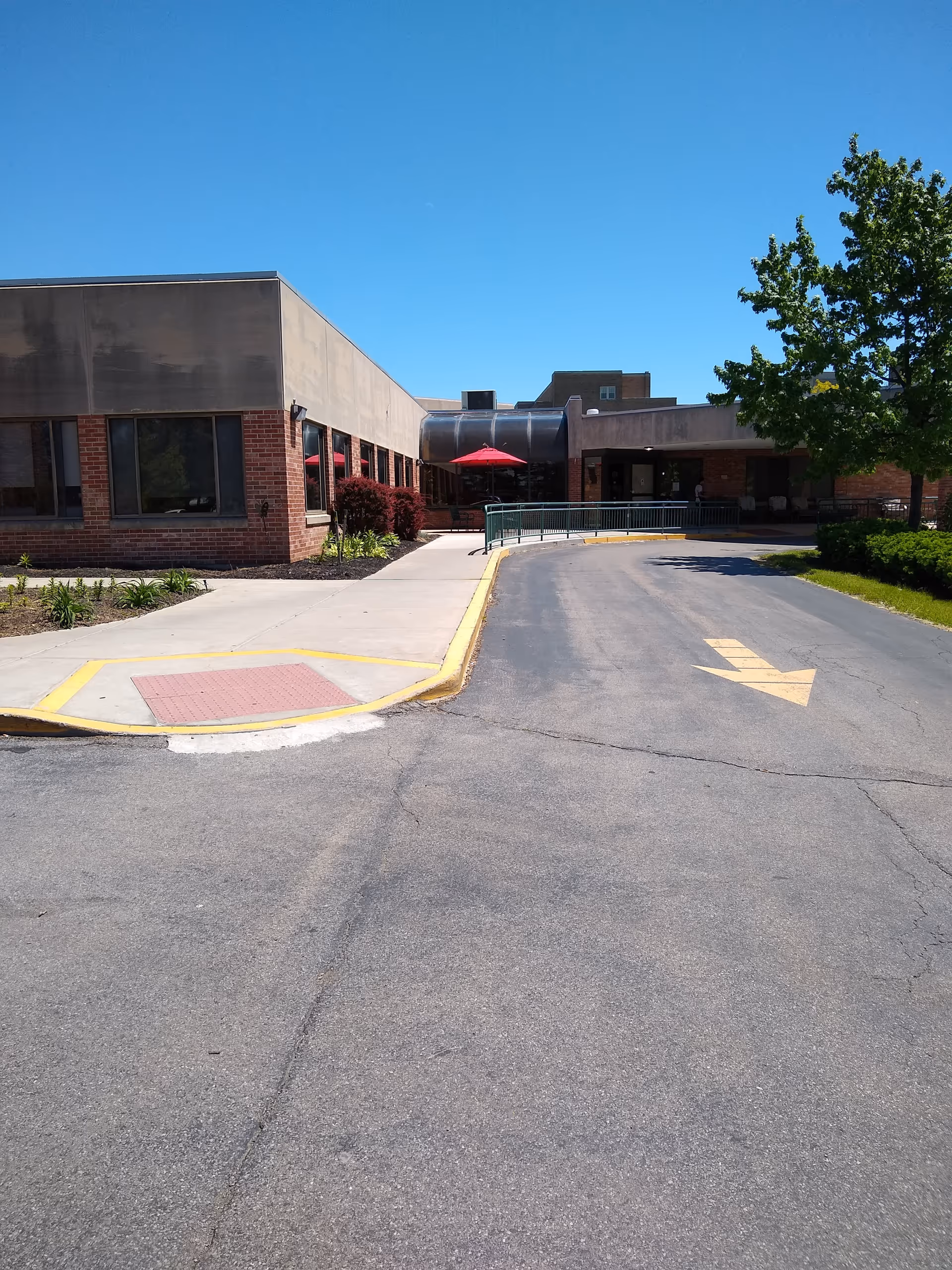 A single-story brick nursing facility exterior with driveway and trees under blue sky.