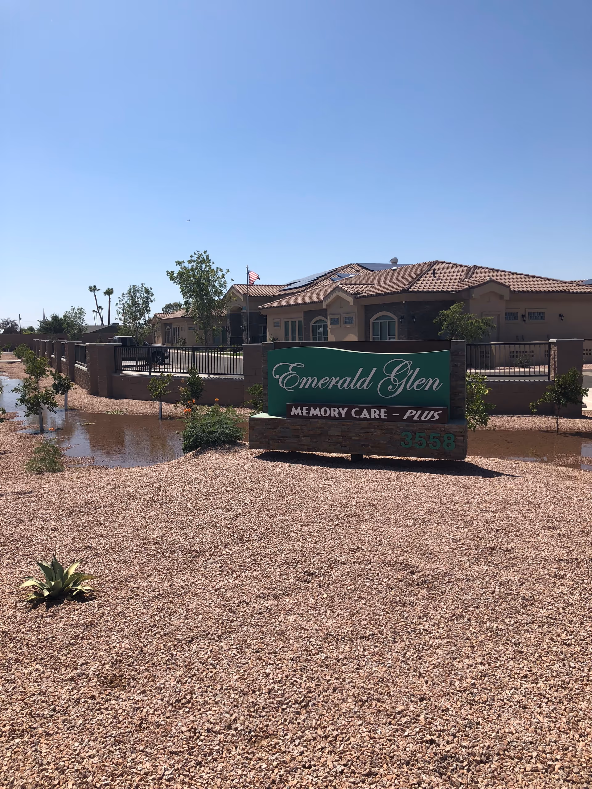 Exterior view of Emerald Glen Assisted Living facility with a green sign that reads 'Emerald Glen Memory Care - Plus' in front of a single-story building with a tiled roof, surrounded by a gravel landscape and some small plants under a clear blue sky.