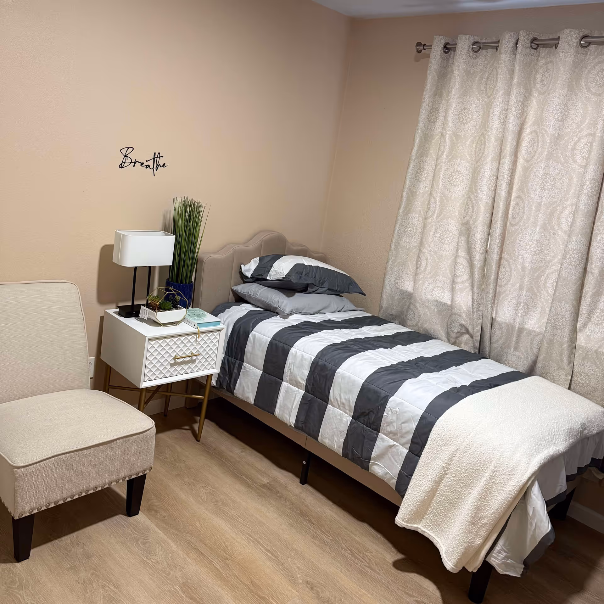 A neat, single bedroom with a bed, nightstand, lamp, chair, and beige walls.
