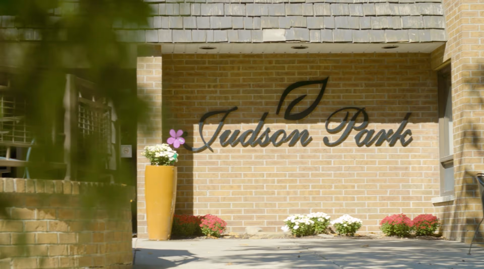 Exterior view of a brick wall with the sign 'Judson Park' mounted on it, accompanied by a leaf logo. There is a tall yellow planter with white flowers and a purple flower decoration next to it, along with small flower beds with white and red flowers at the base of the wall.