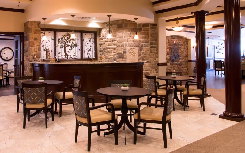 A cozy common area in a senior living facility featuring round tables with four cushioned chairs each, small potted plants on the tables, stone accent walls, and warm lighting fixtures hanging from the ceiling. The space has a welcoming atmosphere with a dark wood reception or bar counter and decorative stained glass windows behind it.
