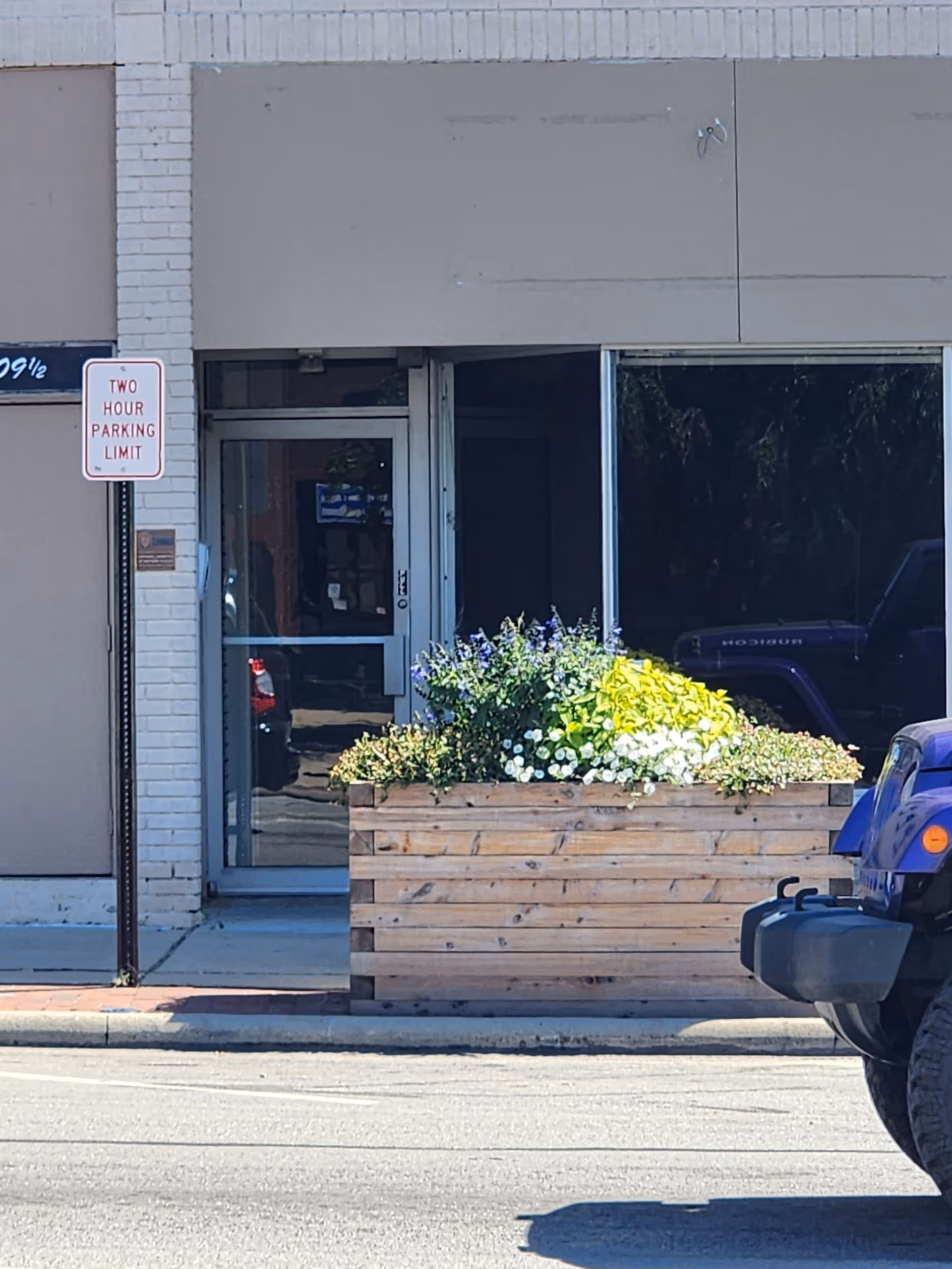 Storefront entrance with glass door, wooden planter filled with flowers, and a two-hour parking sign.