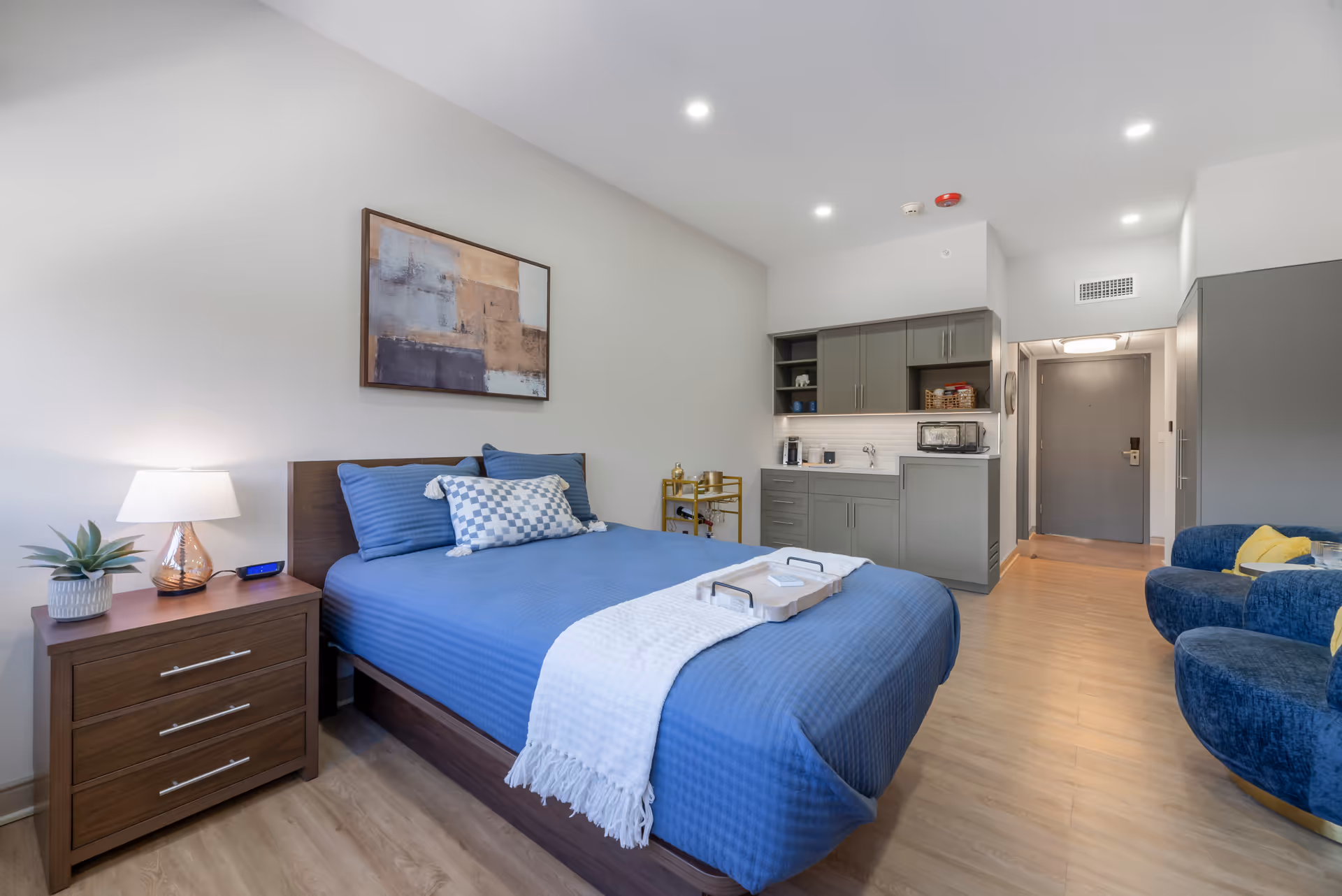 Modern senior living suite featuring a blue bed, kitchenette, and cozy seating area.