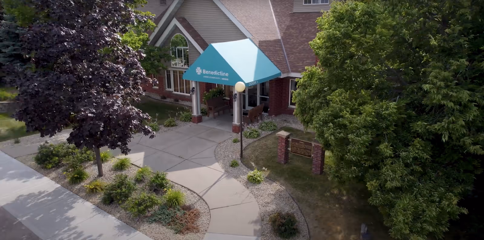 Front entrance of Benedictine Living Community-Osseo with a teal canopy over a walkway and surrounding trees and landscaping.