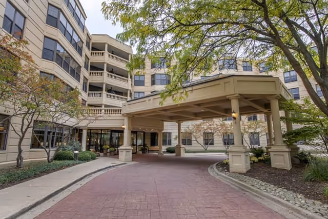 Exterior view of a multi-story senior living facility with a covered driveway entrance, surrounded by trees and landscaping.