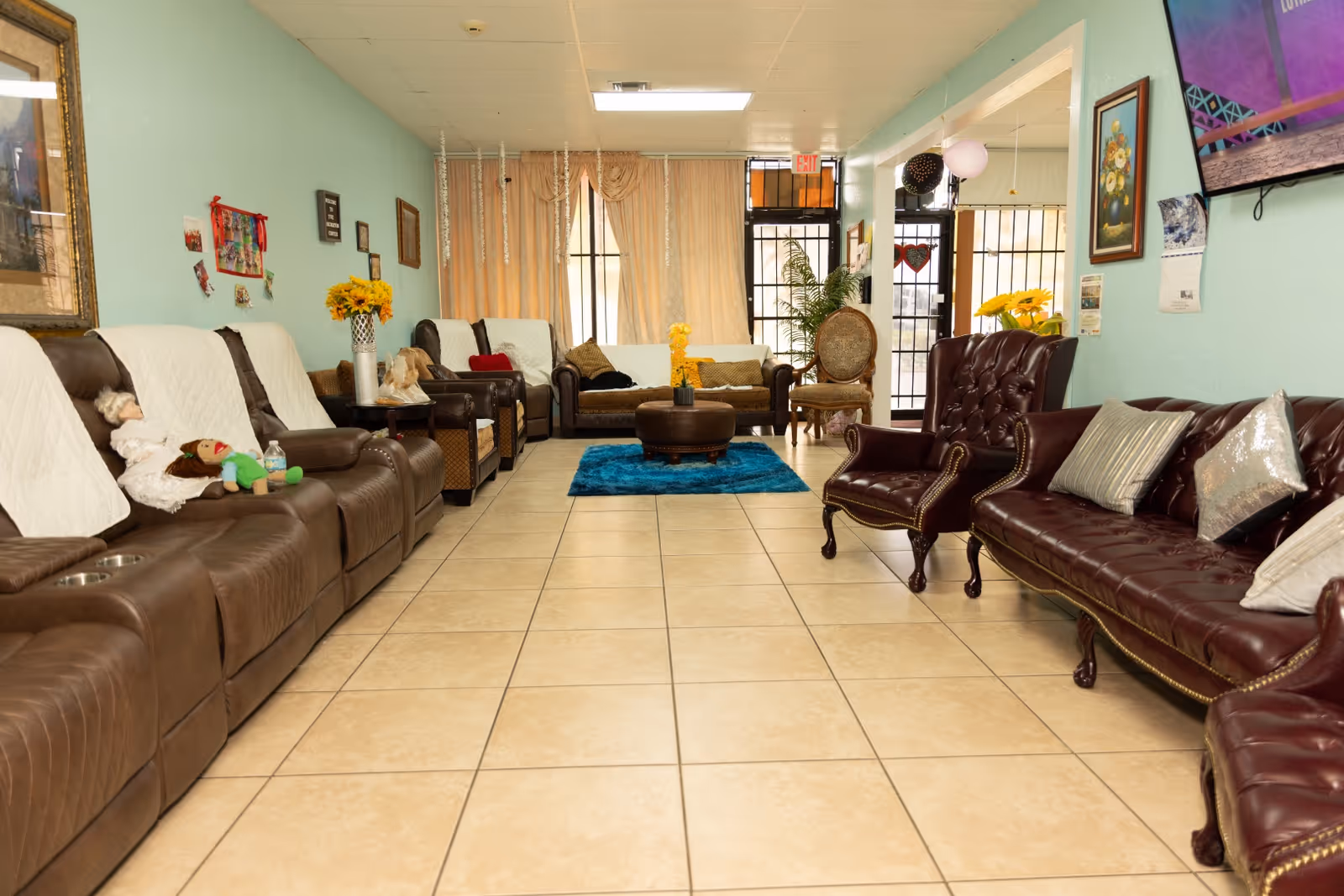 A cozy adult daycare common area with brown leather recliners and sofas arranged along the walls. The room has light blue walls, tiled floor, framed pictures, and decorative flowers on side tables. A blue rug and ottoman are centered in the room, with large windows covered by beige curtains letting in natural light.