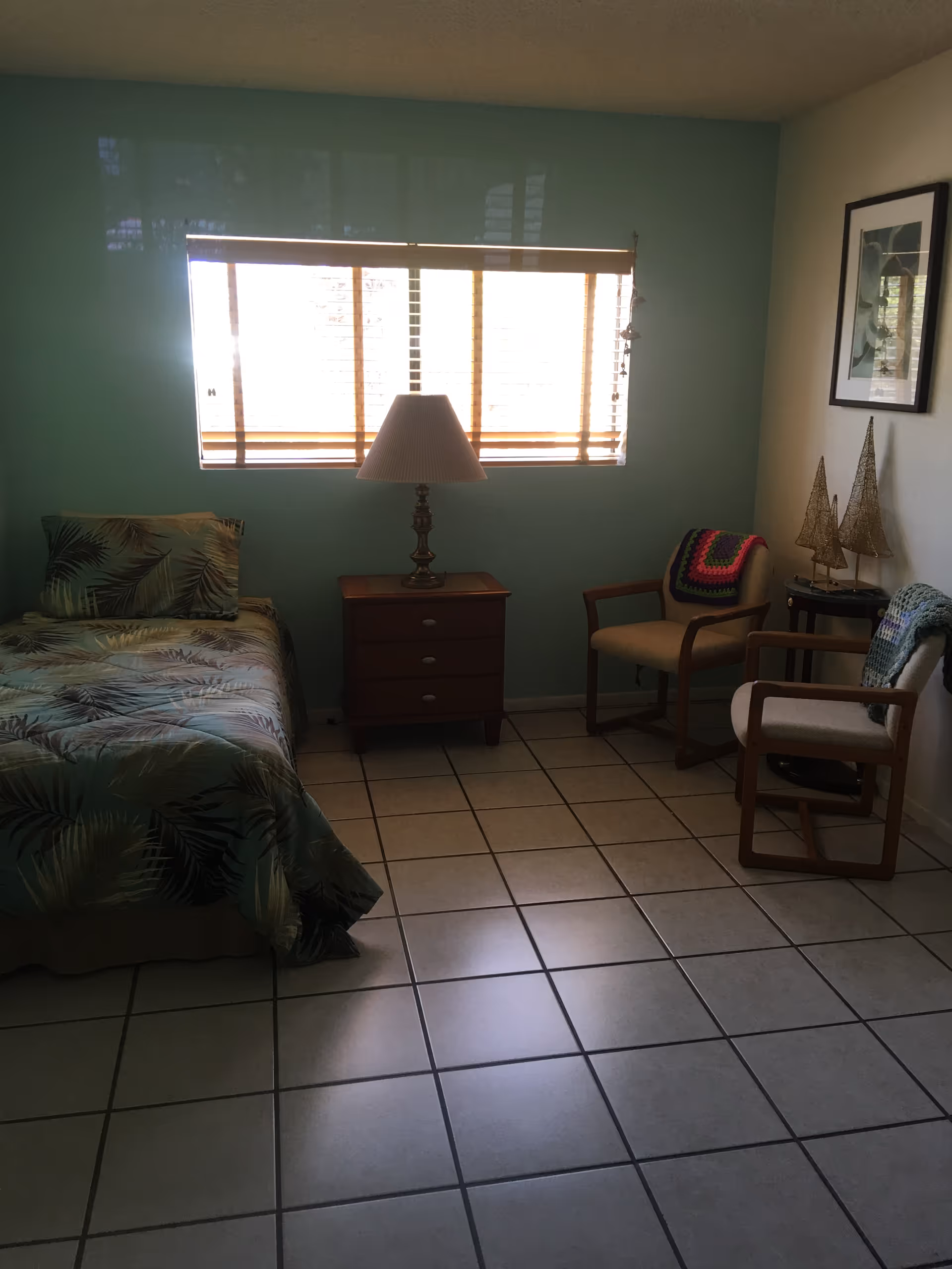 A small turquoise bedroom with a bed, dresser, lamp, window blinds, and chairs.