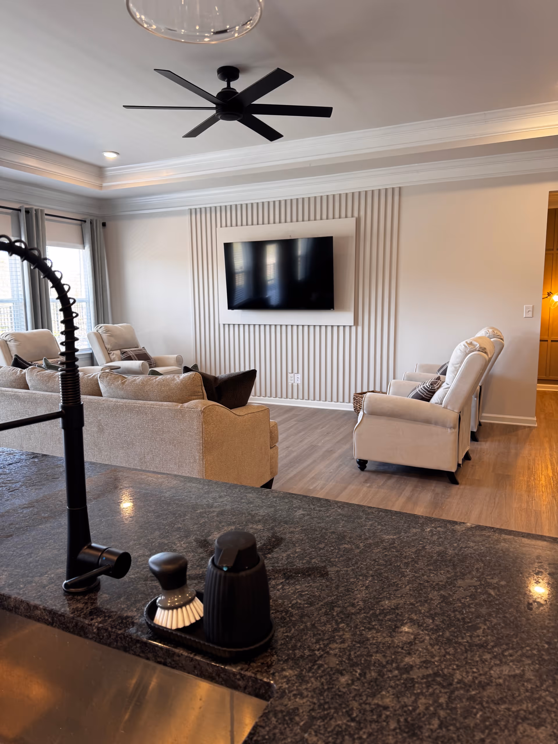 Light-filled living room with beige sofas, a wall-mounted TV, and a dark granite counter in foreground.
