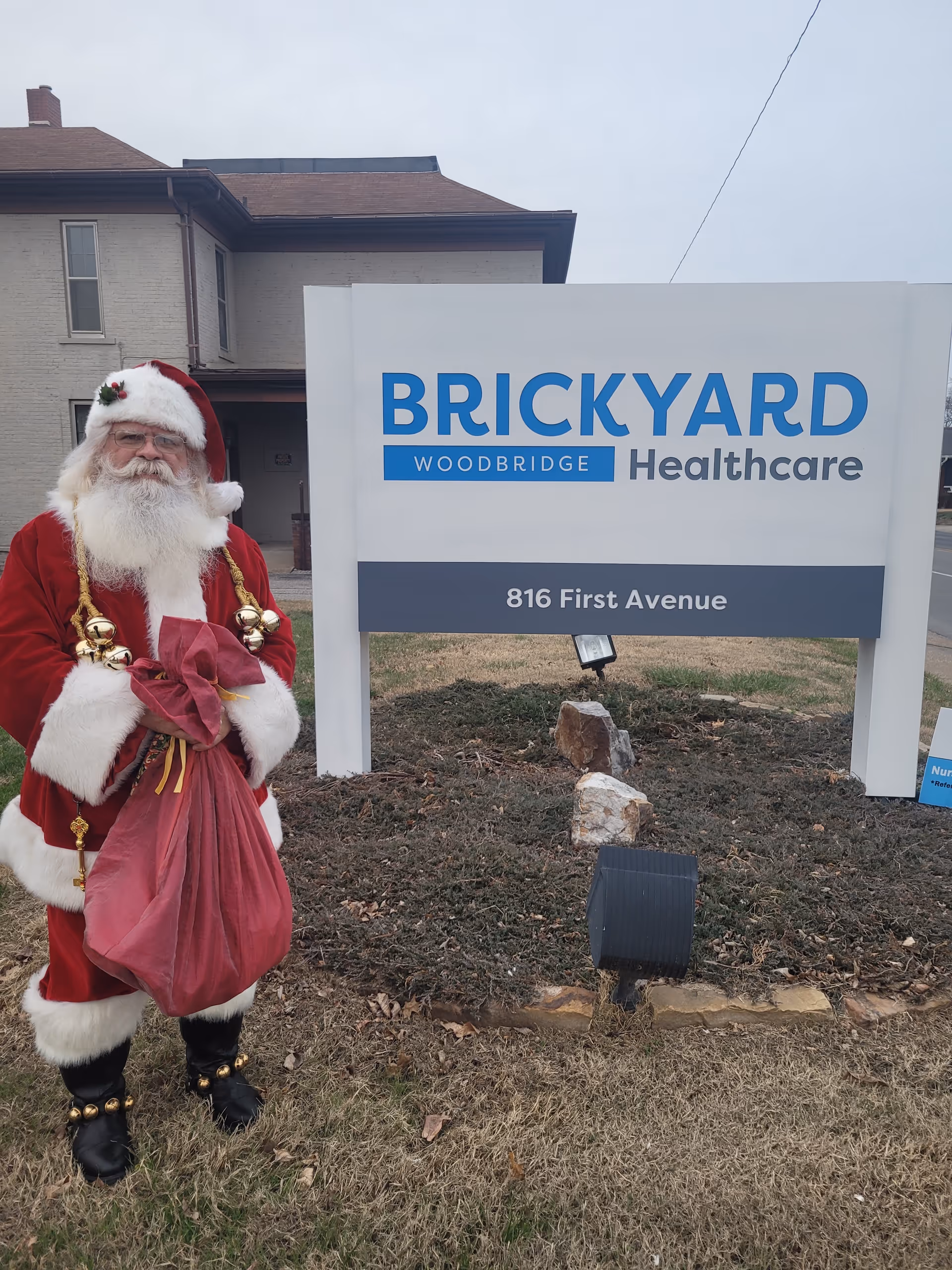 Santa stands outside a Brickyard Woodbridge Healthcare sign near a building.