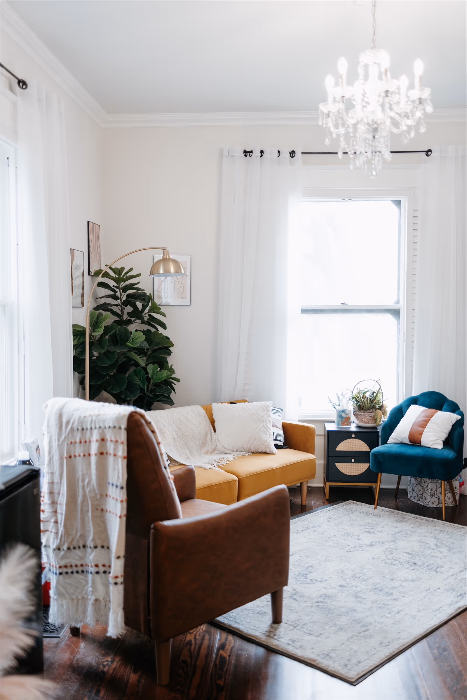 Bright living room with mustard sofa, teal chair, chandelier, plants, and a large window.