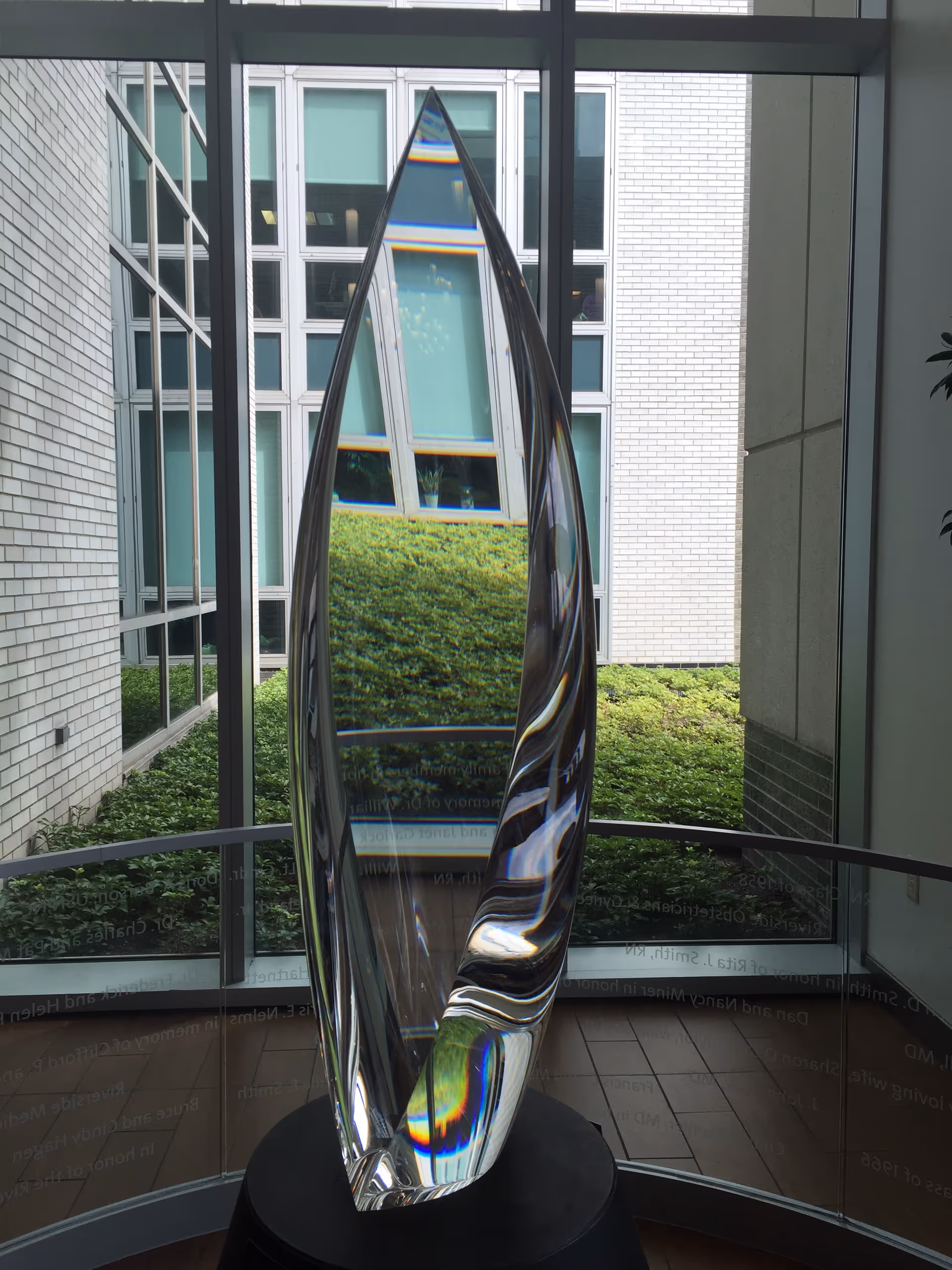 A tall glass sculpture stands in a modern interior atrium with large windows and greenery outside.