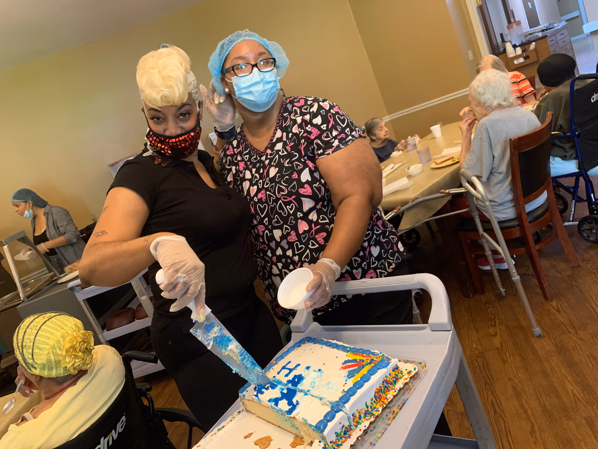 Two masked staff members serve cake to seated residents in a dining room of an assisted living facility.