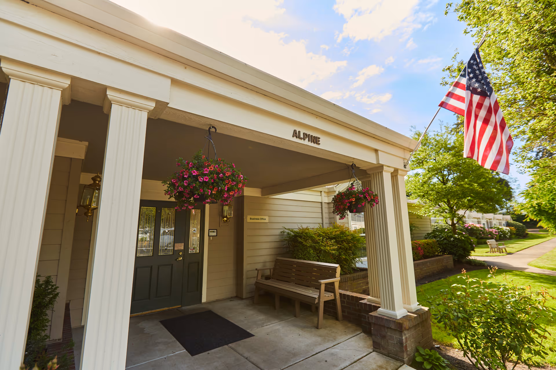 Entrance to a building labeled 'ALPINE' with a covered porch supported by white columns, hanging flower baskets, a wooden bench, and an American flag flying on a pole. The surrounding area has green trees, bushes, and a sidewalk.