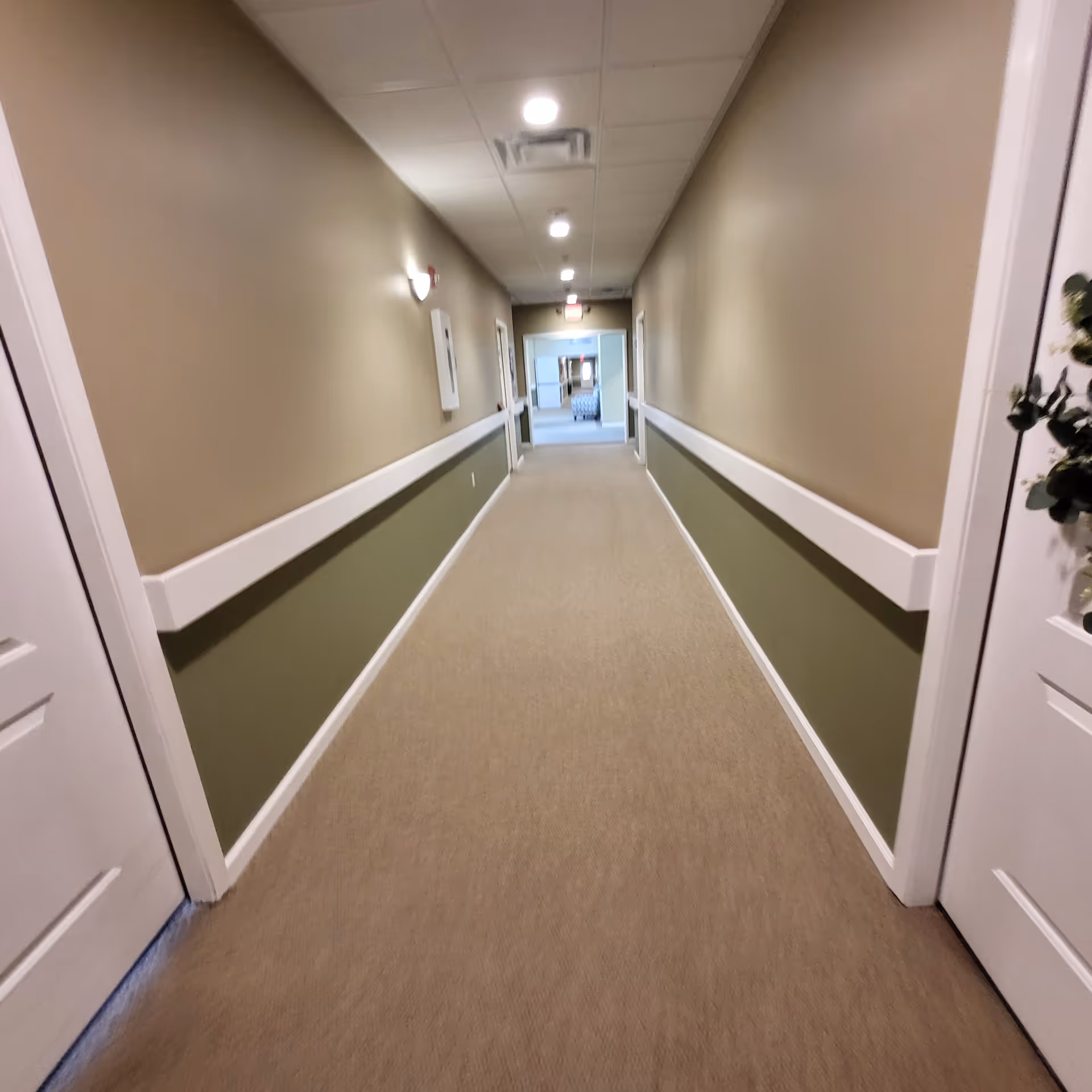 Long indoor hallway with beige walls, white trim, and carpeted floor in a senior living facility.