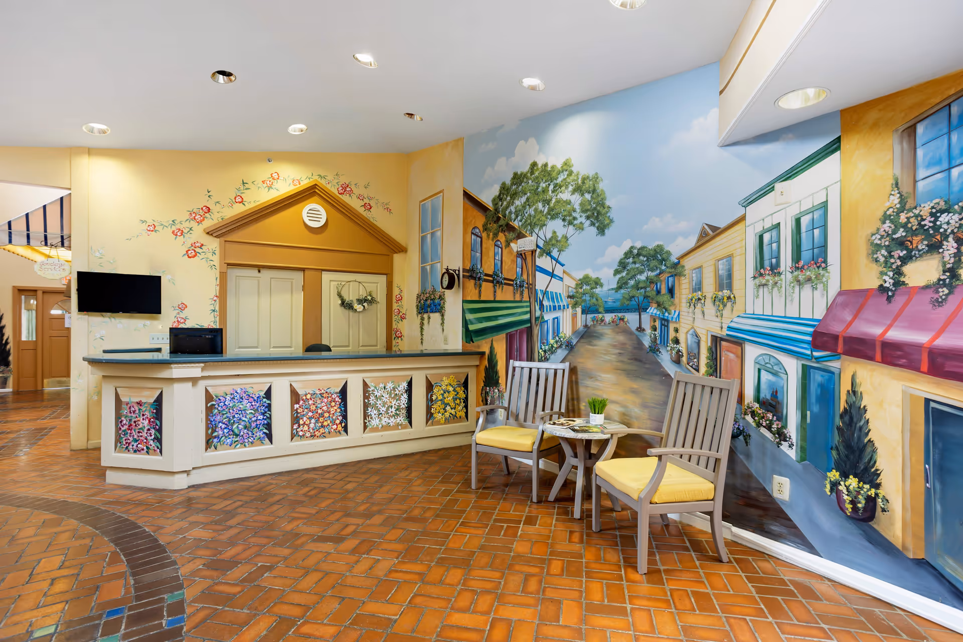 Lobby reception area with a decorative front desk, two chairs and a small table in front of a large street-scene mural.