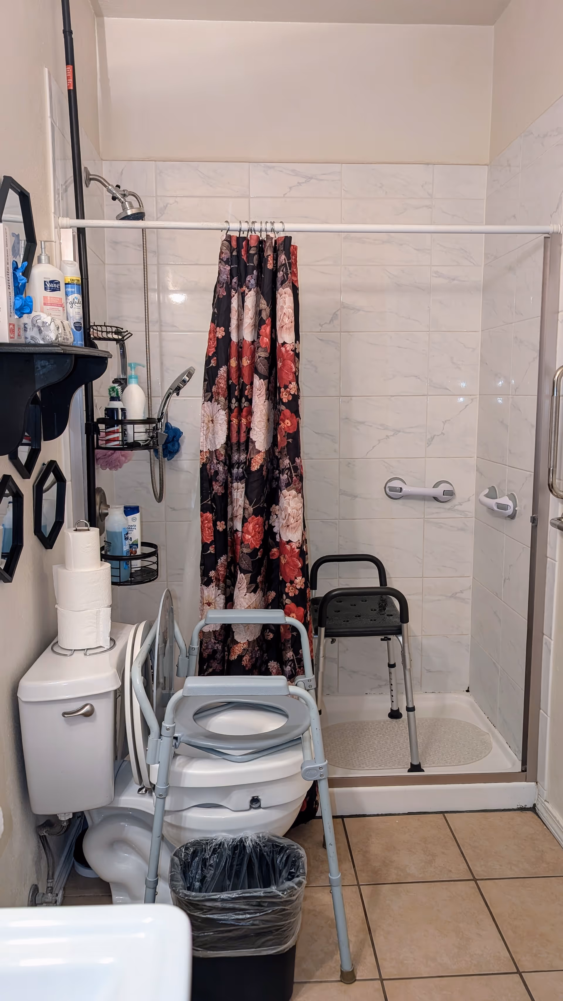 Accessible bathroom with floral shower curtain, shower chair, walker, toilet, and stocked shelves.