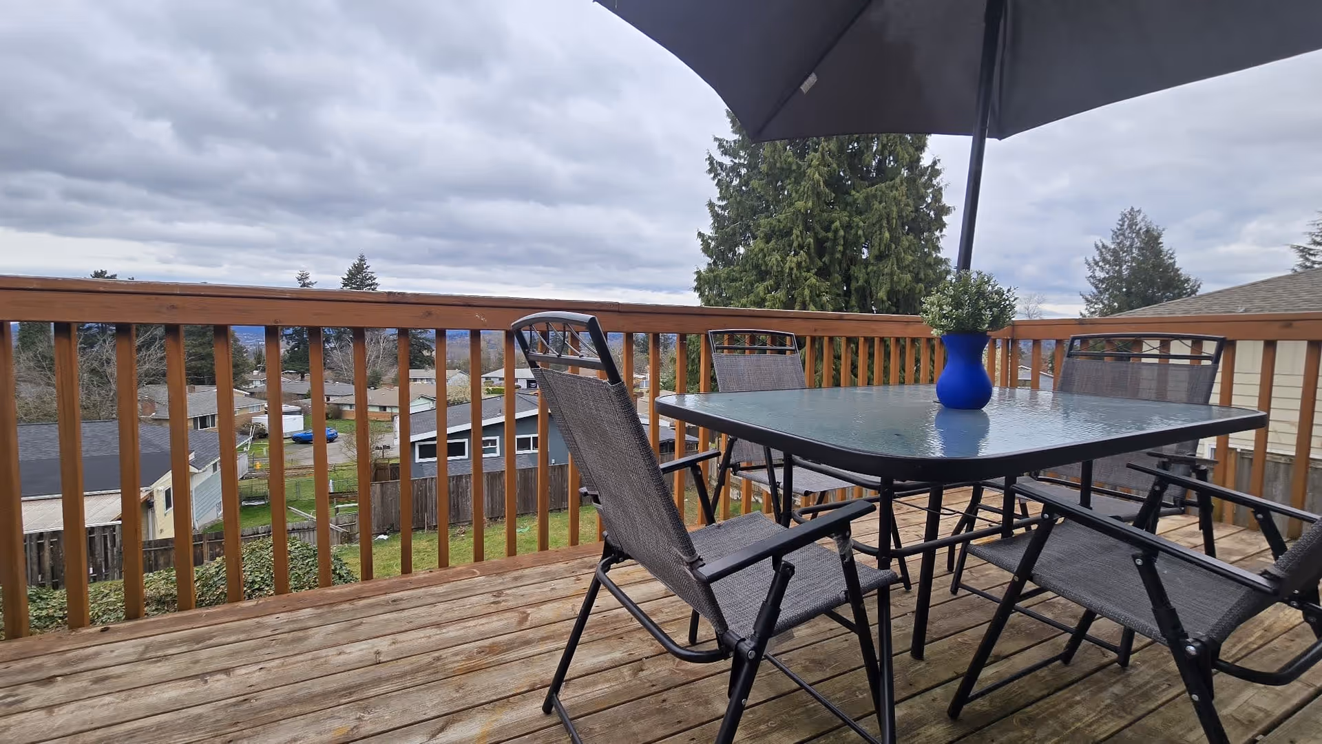 Outdoor wooden deck with glass table, chairs, umbrella, blue vase, railing, and distant neighborhood homes.