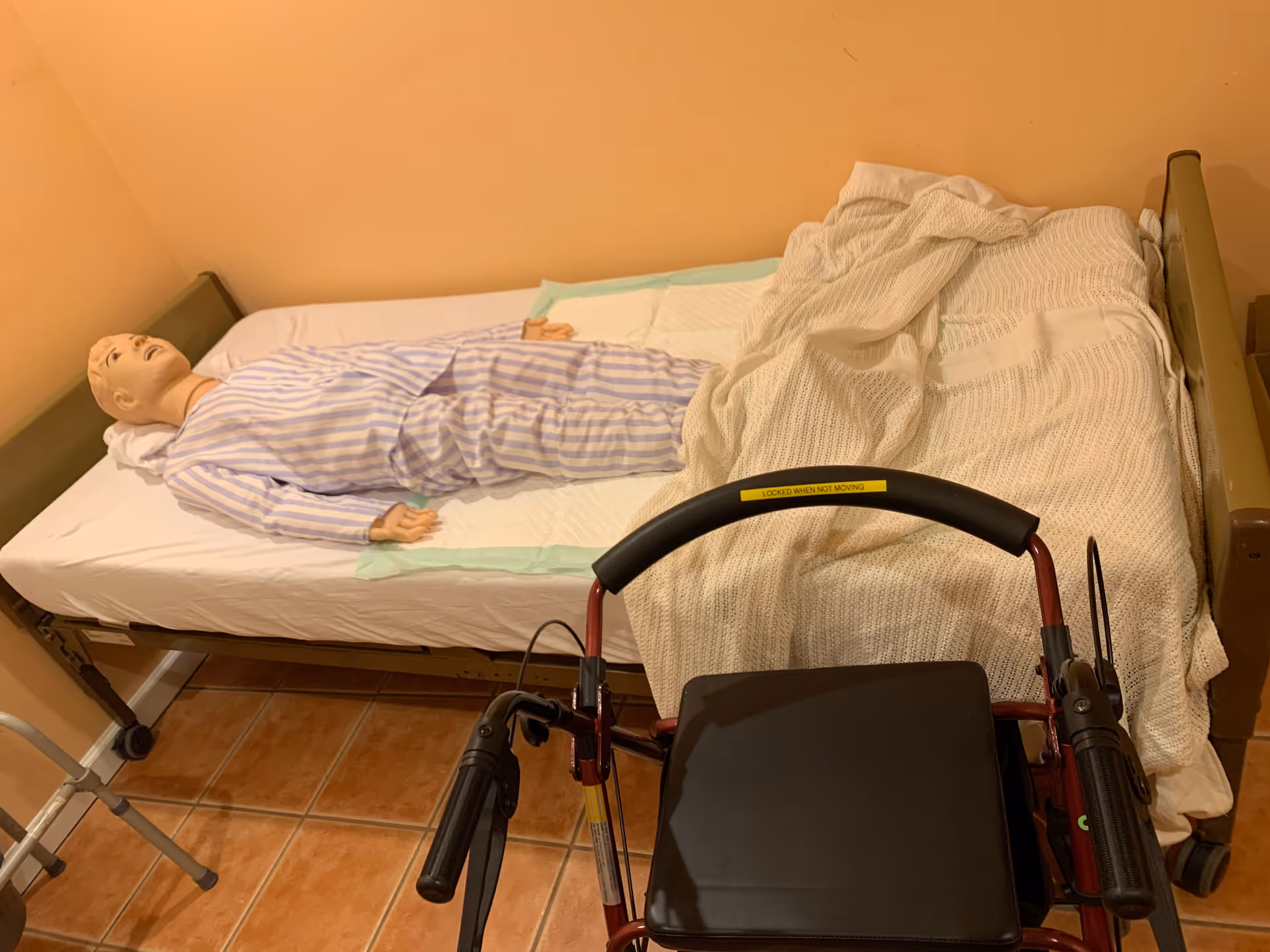 A medical training mannequin lies in a hospital-style bed beside a wheelchair in an interior room.