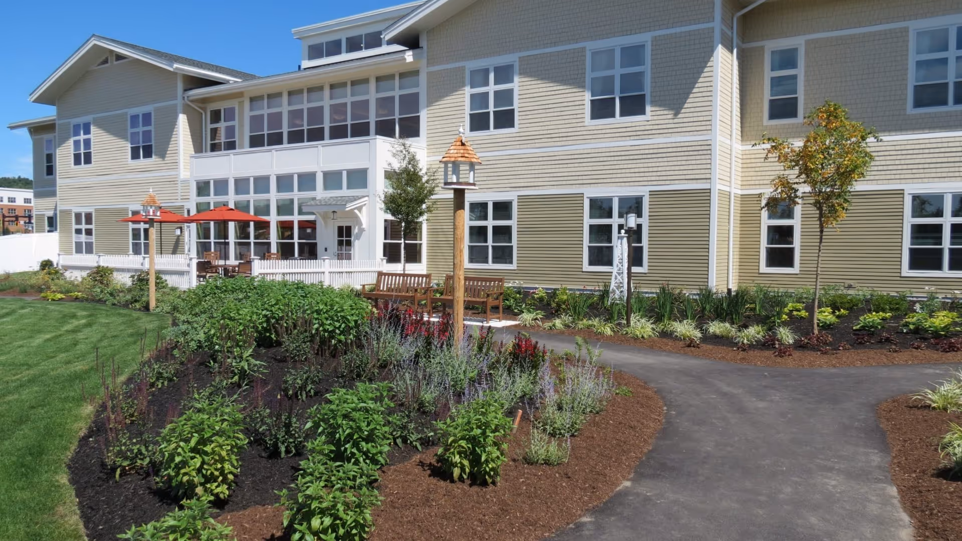 Exterior view of a two-story beige senior living building with landscaped gardens, pathways, benches and patio umbrellas.